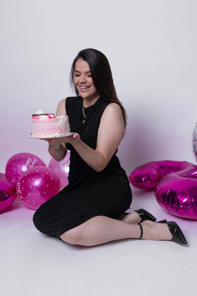Joyful woman in black dress celebrating birthday with balloons and cake indoors.